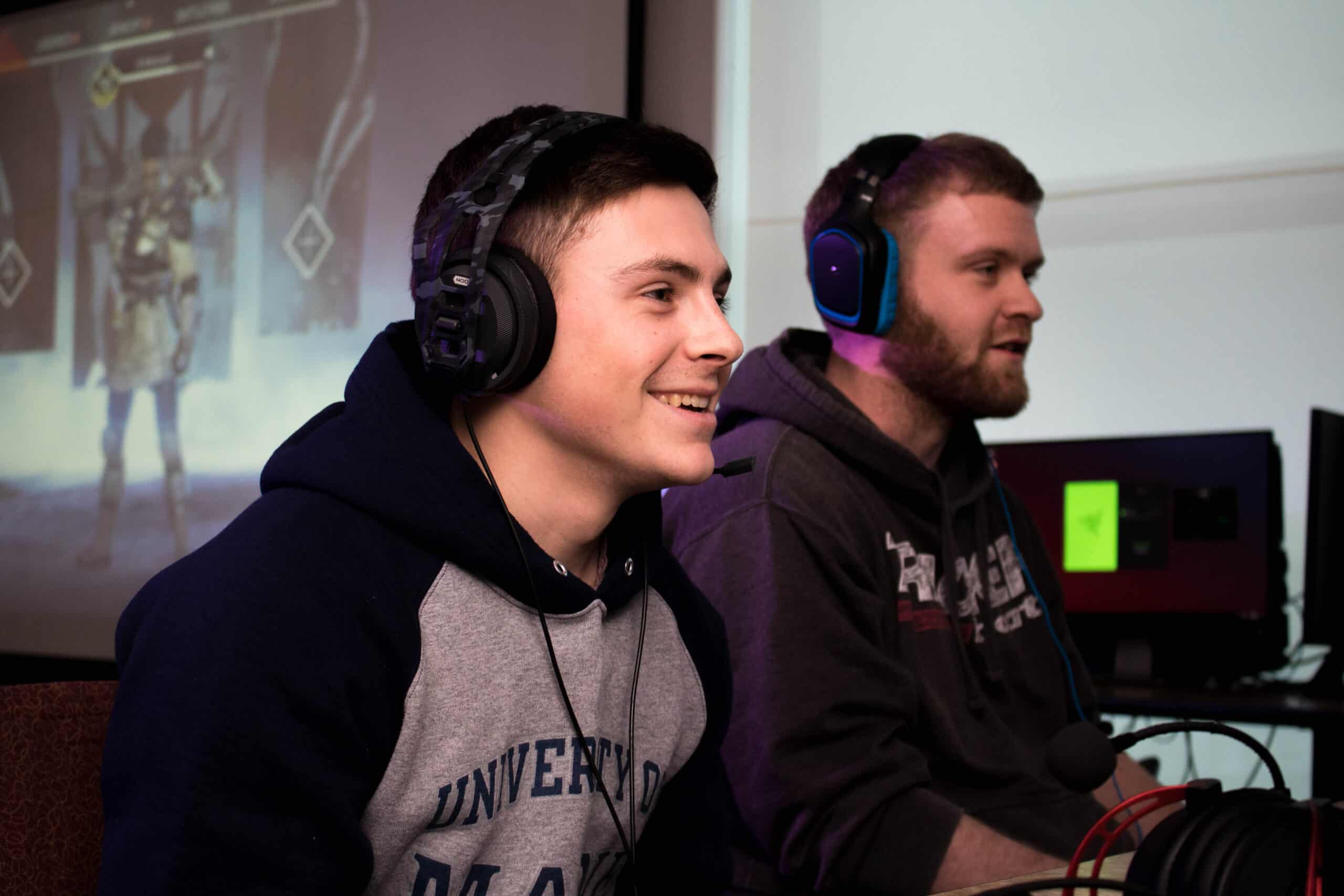 Two young men wearing headphones, smile at a computer that is off screen, They seem to be playing video games.
