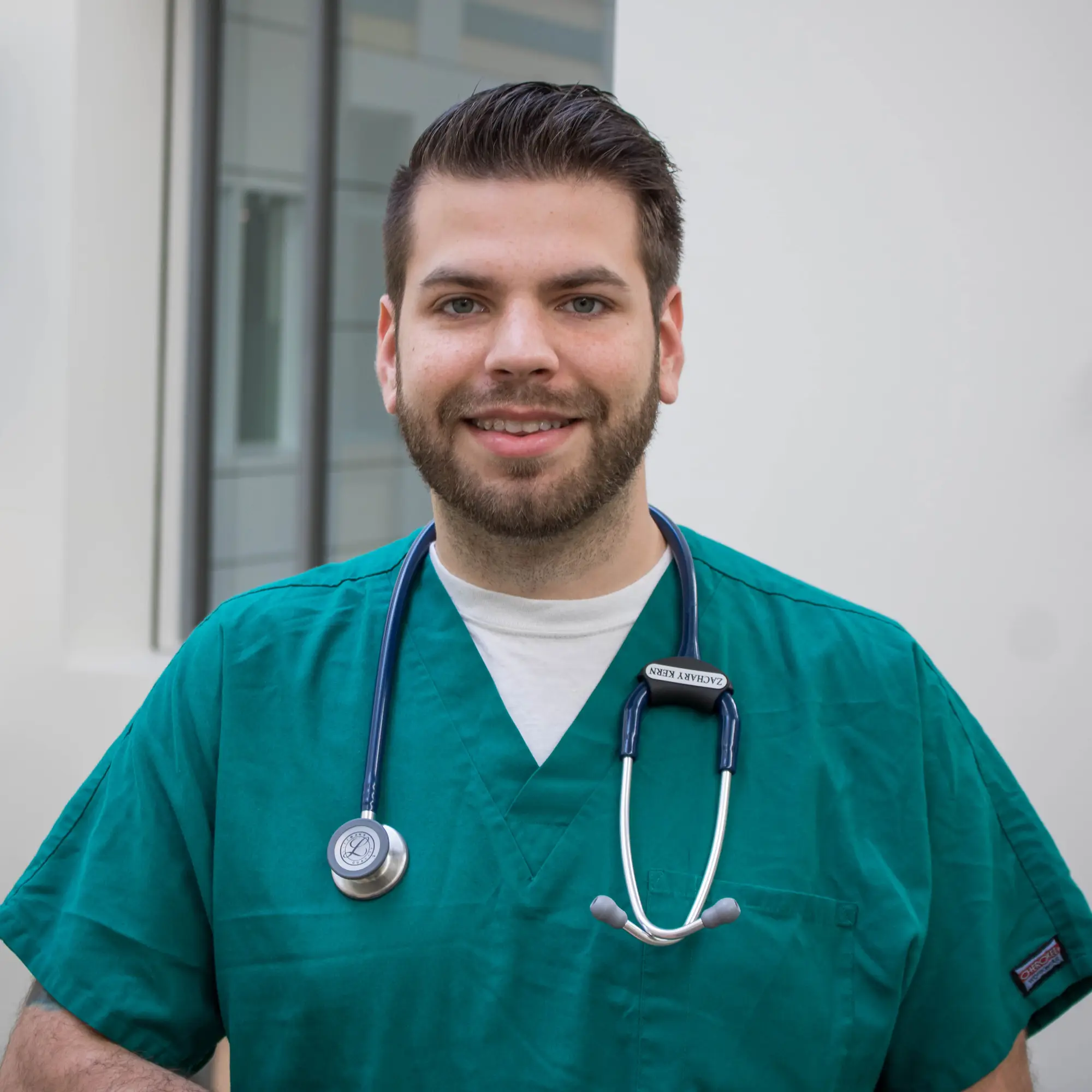 A man with short hair and a beard wearing scrubs and a stethoscope smiles at the camera.