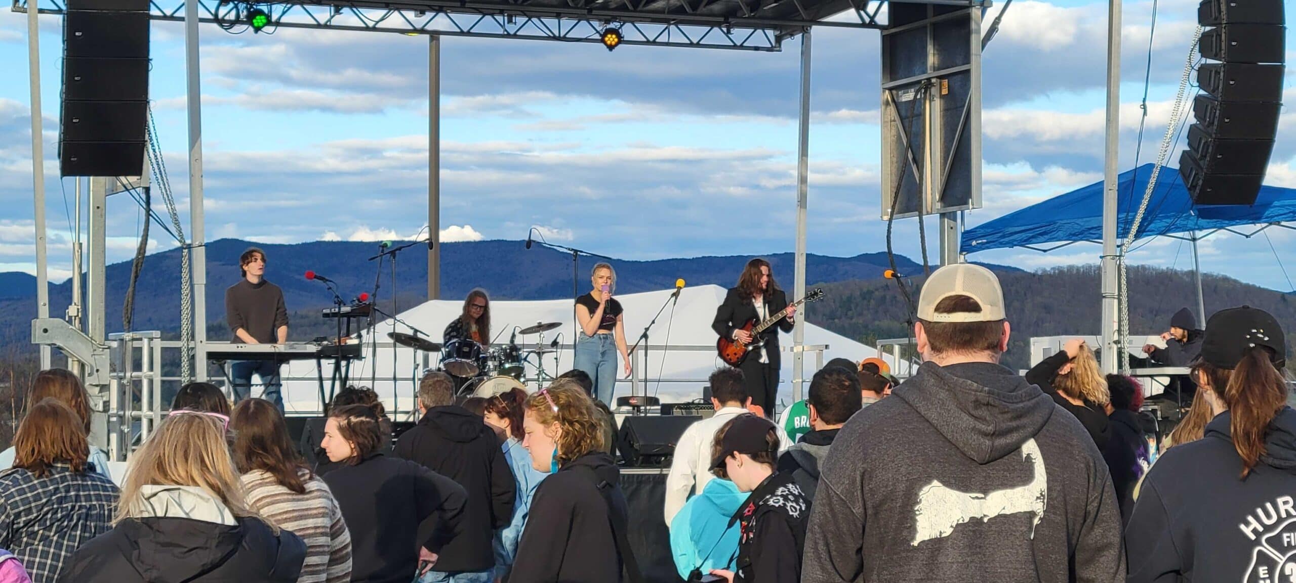 A band on a stage with mountains behind them as a crowd listens in front of them.