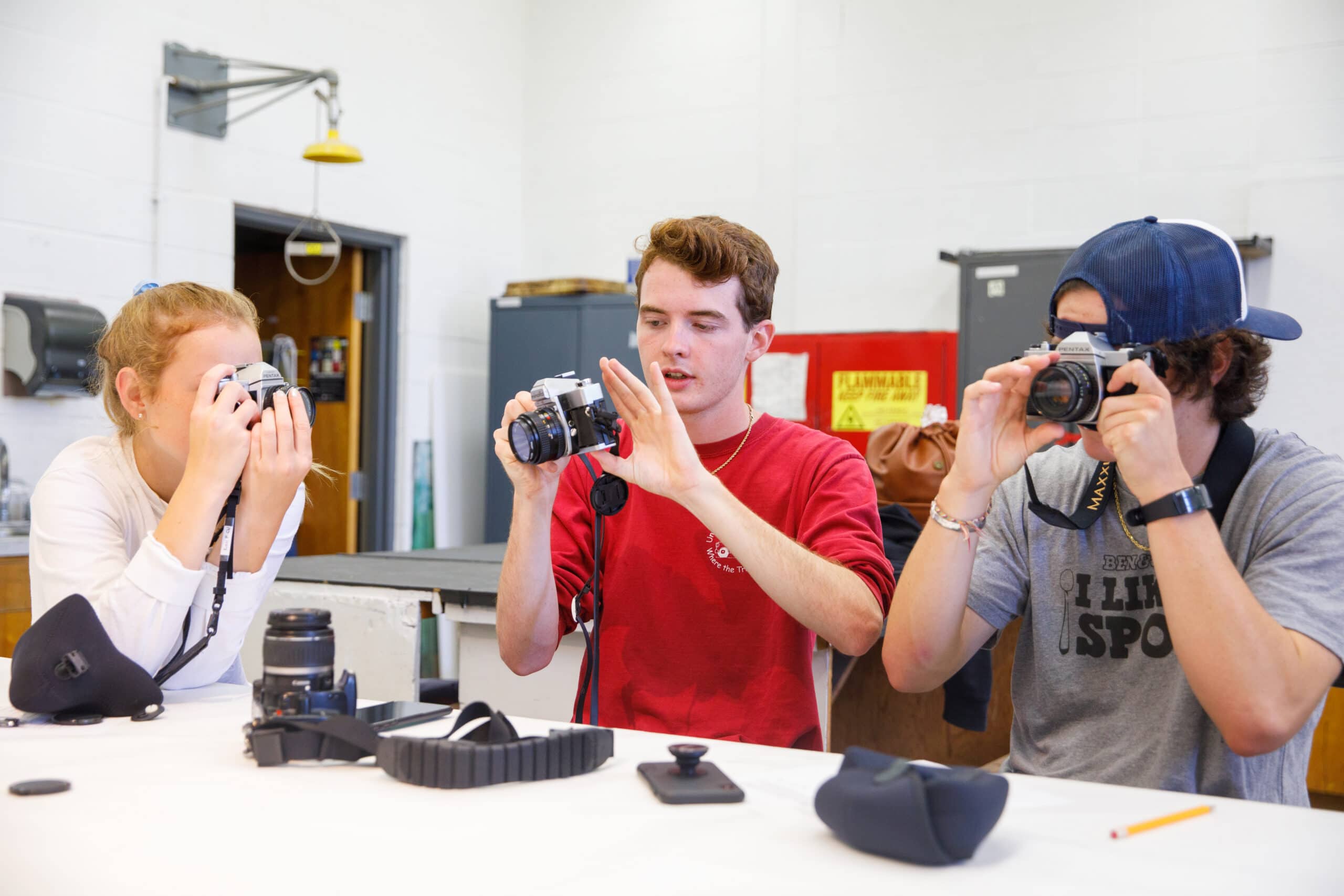 Two young guys and a girl are playing with camera equipment while leaning on a desk.
