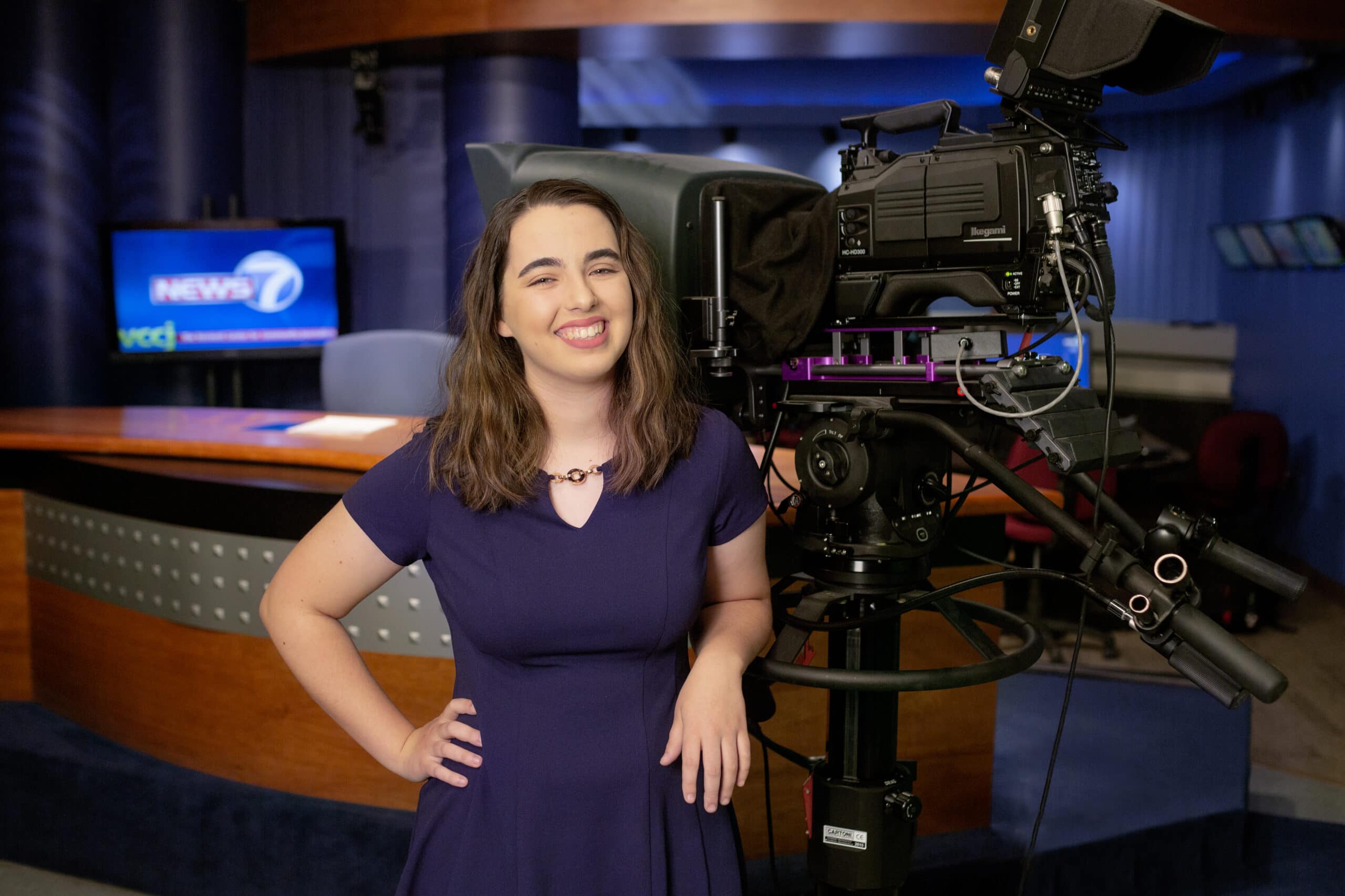 A young woman with long brown hair wears a purple dress while standing in a news studio leaning on a camera.