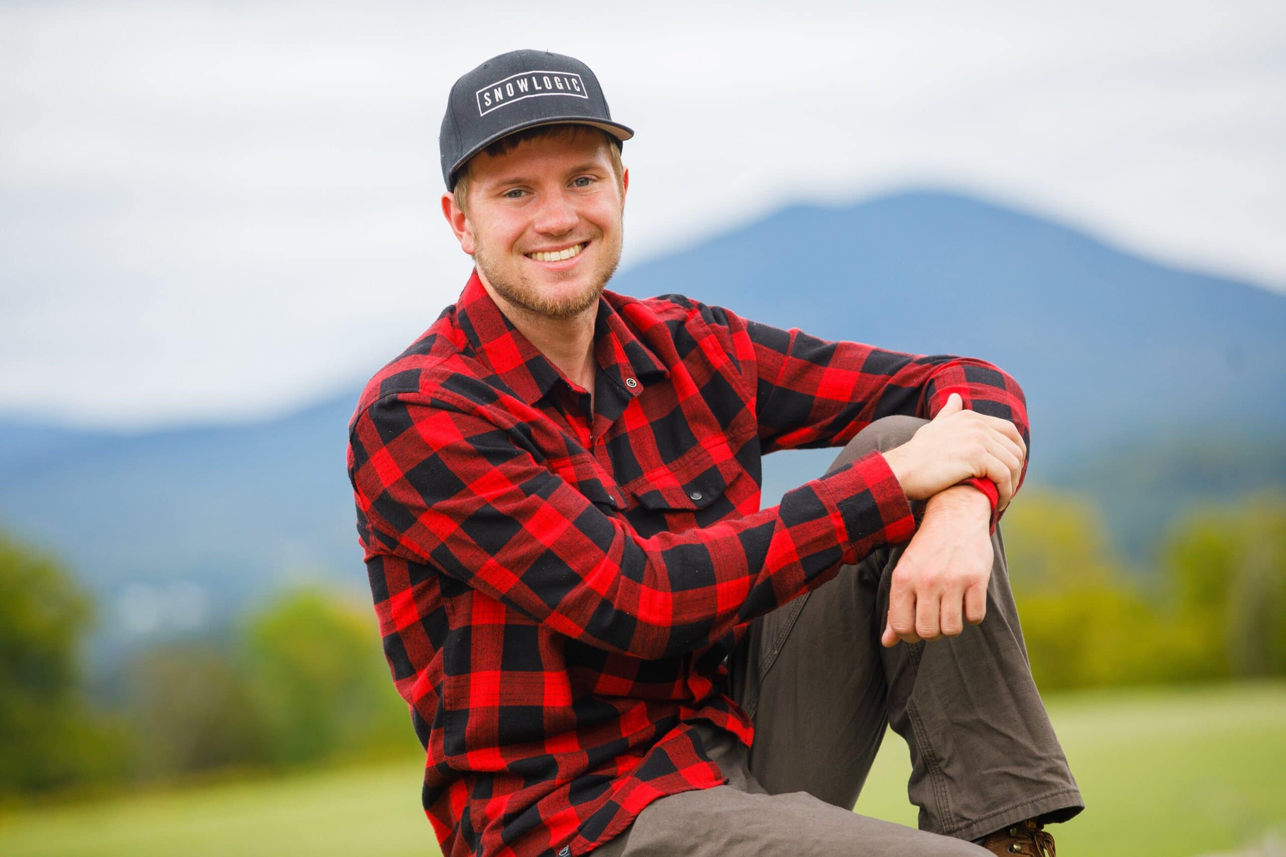 A young man in a baseball cap wearing a red check flannel shirt smiles at the camera.