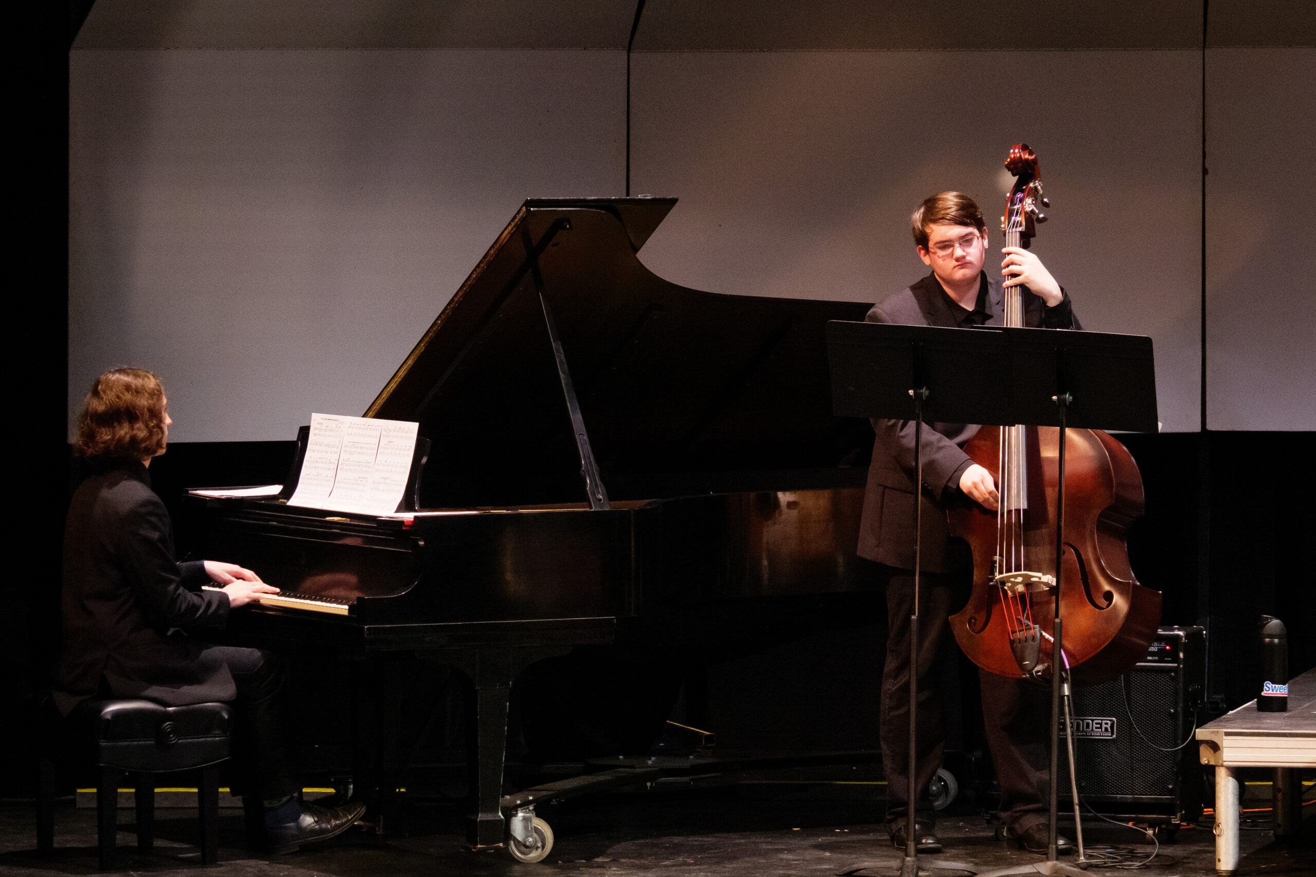 Two student performing on stage, one sitting at a grand piano and another standing and playing an upright bass