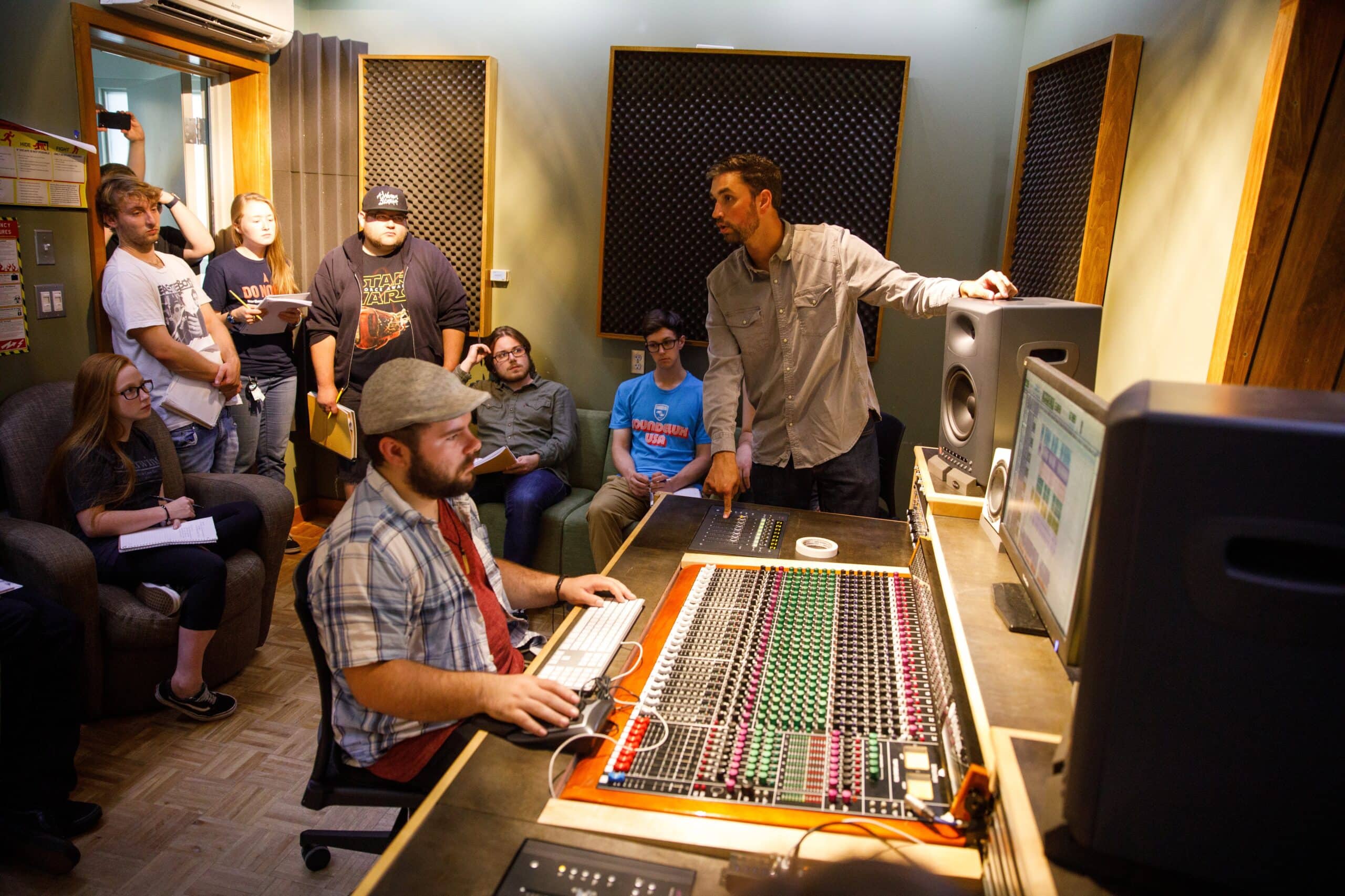 A group of people stand in a recording studio watching one young man run an audio mixer.