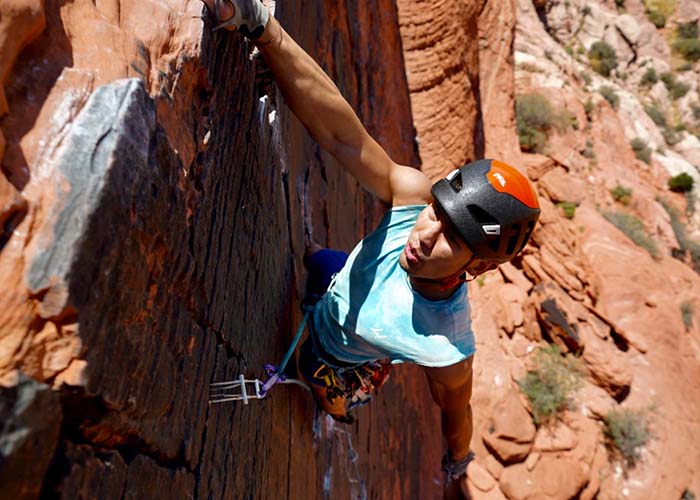 A guy wearing a helmet hangs by one arm from the edge of a desert cliff.