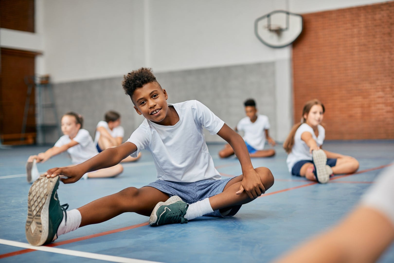 Happy black elementary student stretching his leg while warming up during physical education class at school gym and looking at camera.