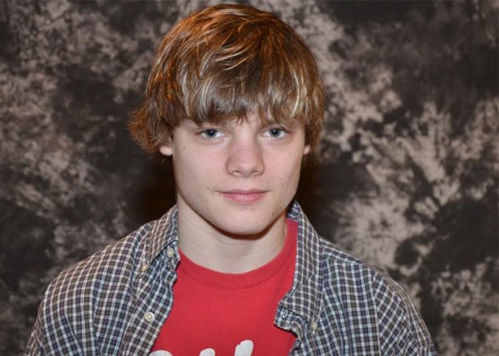 A young man with shaggy blond hair wearing a checkered shirt smirks at the camera.