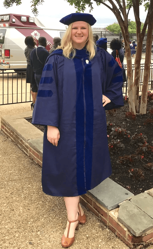 a woman in a graduation gown standing in front of a tree.