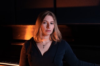 A young woman with shoulder length blond hair leans on a piano.