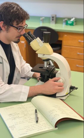 A young man in a lab coat and glasses sits at a desk with a microscope.