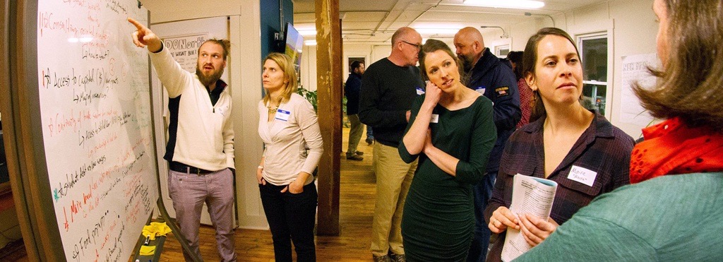 A group of people stand around a white board discussing its content.