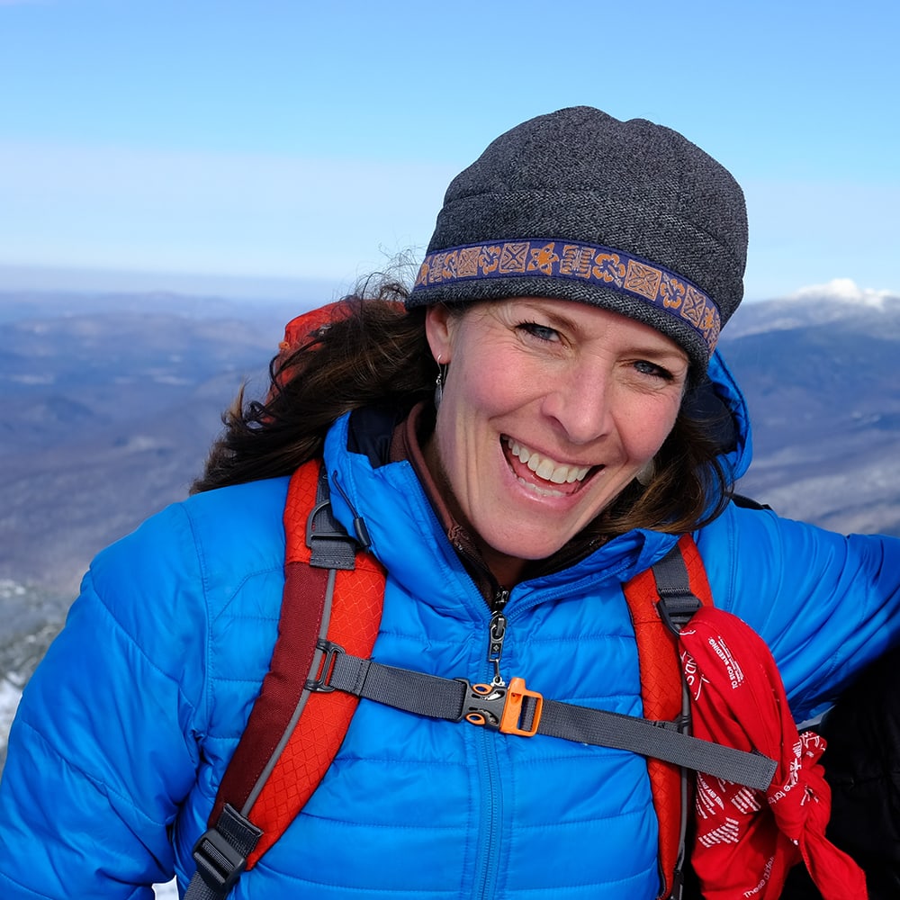 A woman in a winter hat and parka smiles at the camera from the top of a mountain.