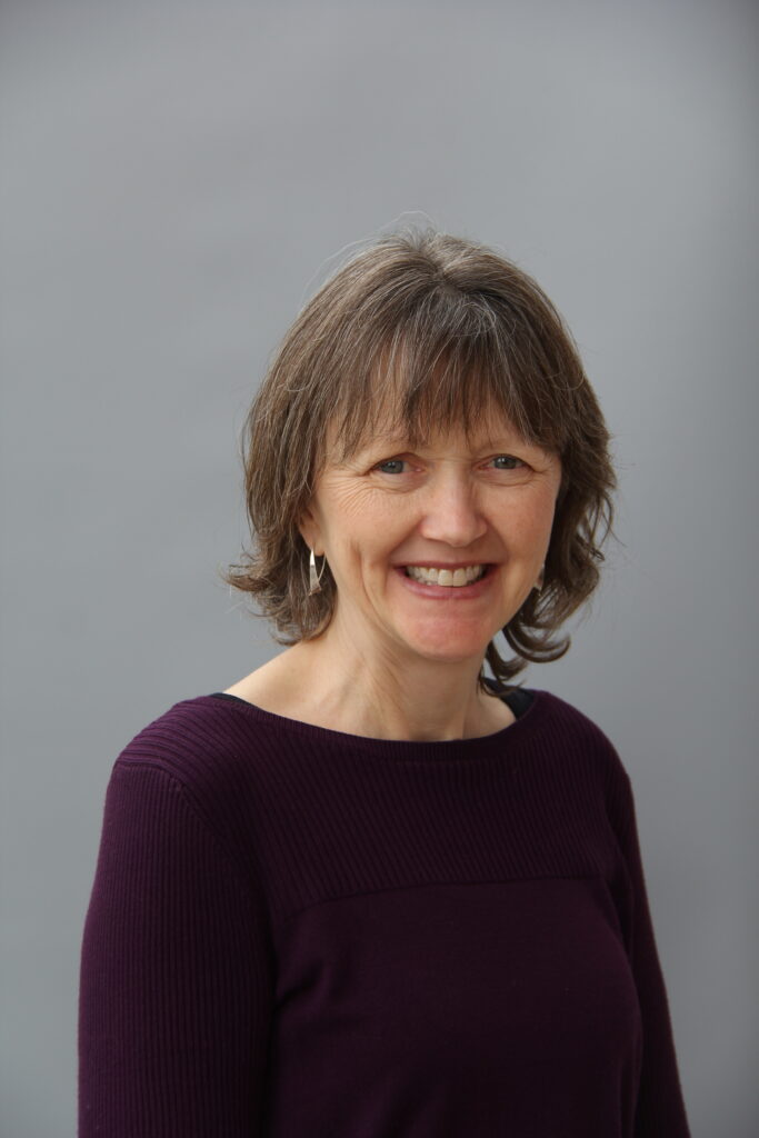 Brown haired women in a brown sweater smiles at the camera, behind her is a grey background.