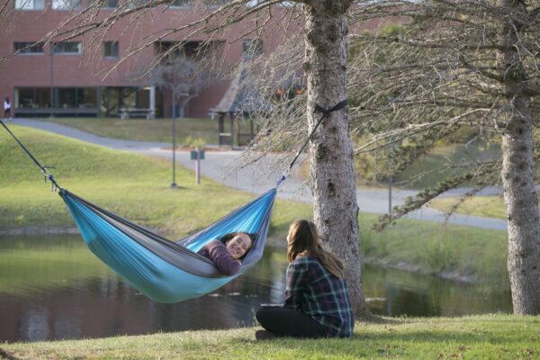 A girl sitting on the ground talking with a girl in a hammock on the Vermont State University Lyndon campus.