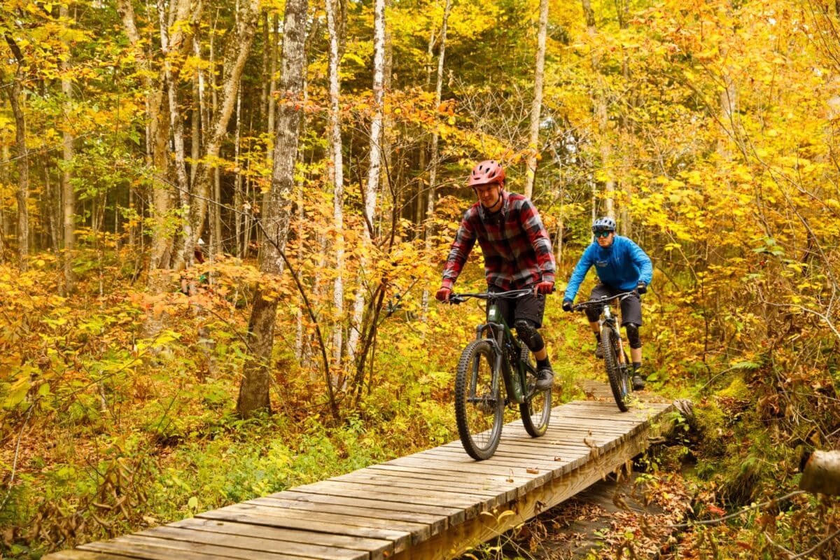 Two bikers cross a bridge in the woods on the Vermont State University Lyndon campus.