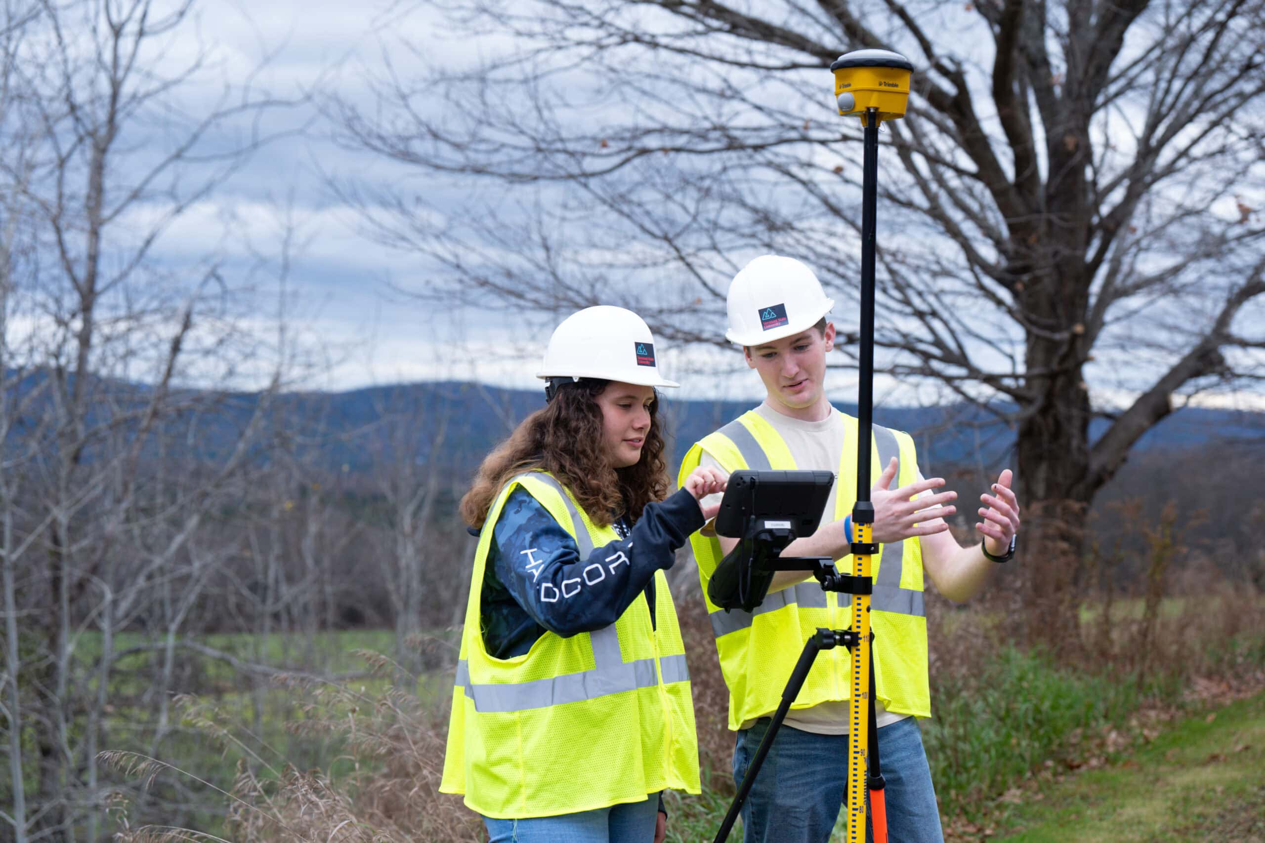 A young man and young woman standing side by side working with a piece of surveying equipment. They're wearing yellow high visibility vests and white hard hats adorned with the VTSU logo in the center.