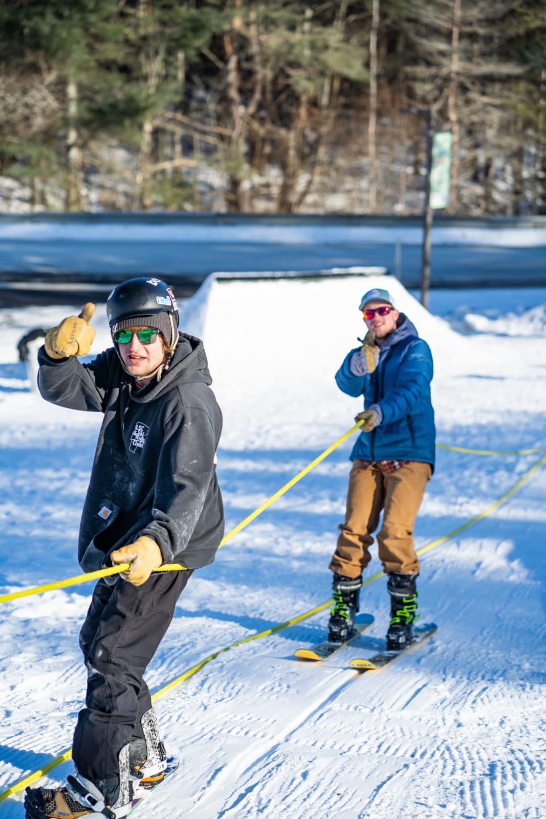 A skier and snowboard give a thumbs-up to the camera on a rope tow at Vermont State University Lyndon.