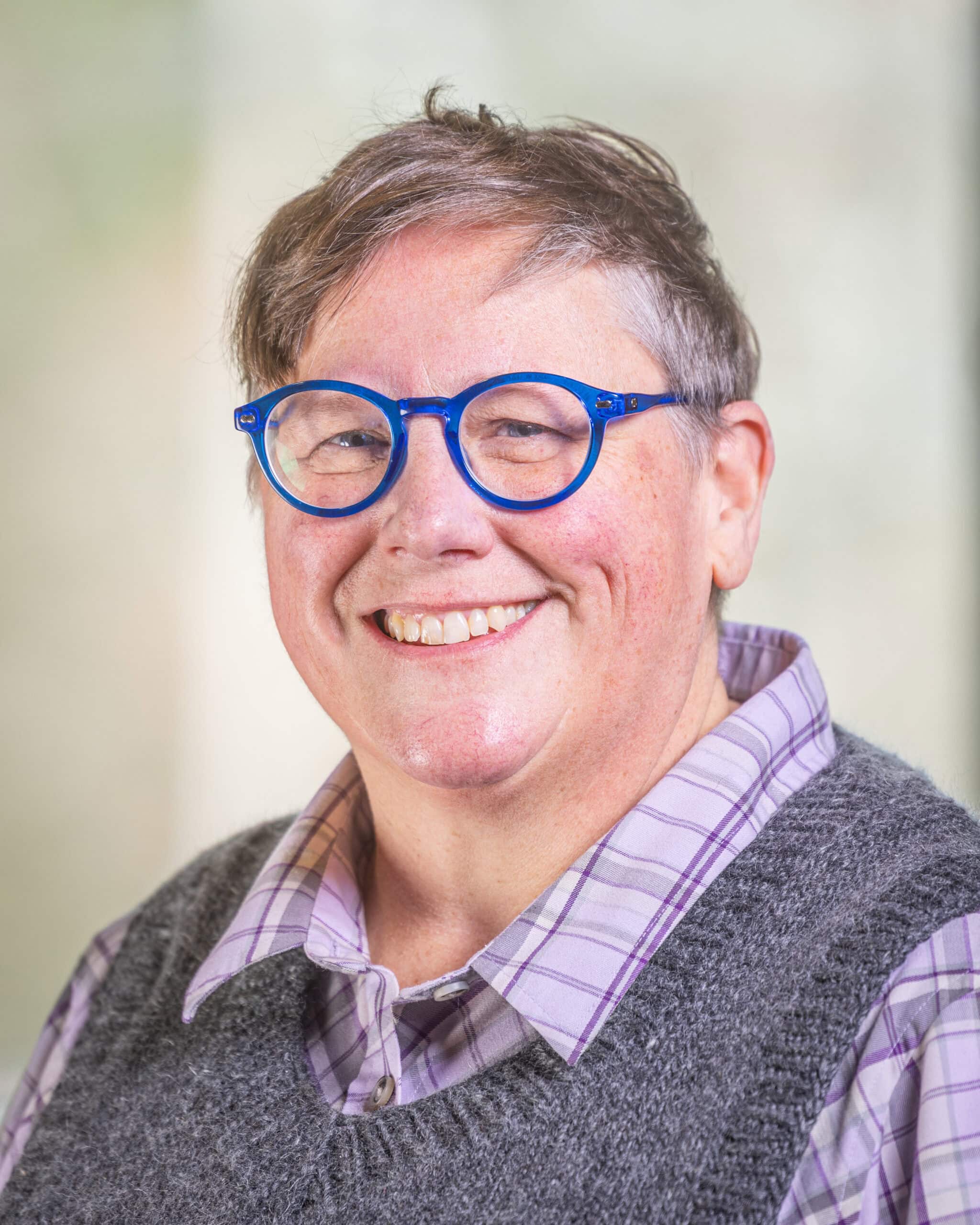 Lisa Zinn, a woman with short brown hair and round blue glasses. She's wearing a purple checkered shirt and a grey sweater-vest and is smiling at the camera.