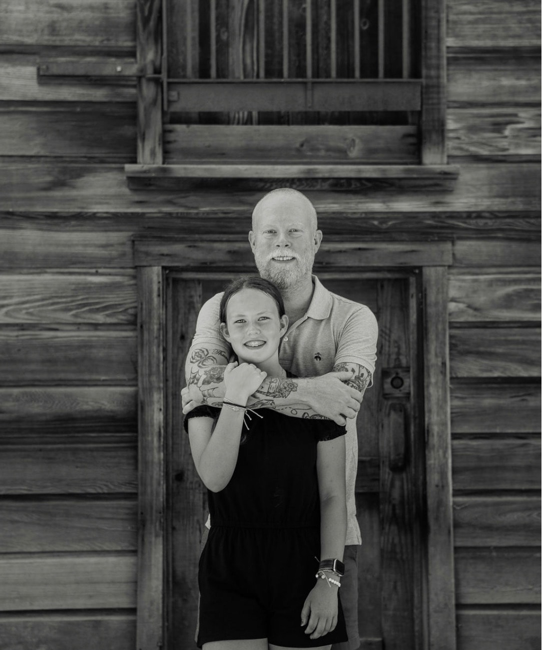 Black and white photo of a man and girl smiling in front of a wooden building.