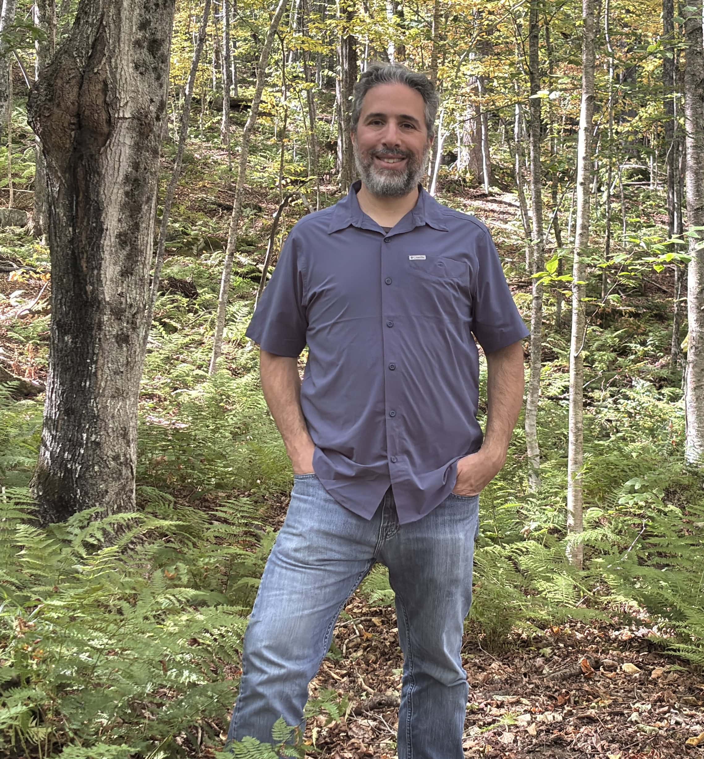 William (Bill) Landesman standing in the woods wearing a grey polo shirt and blue jeans. He's smiling at the camera.