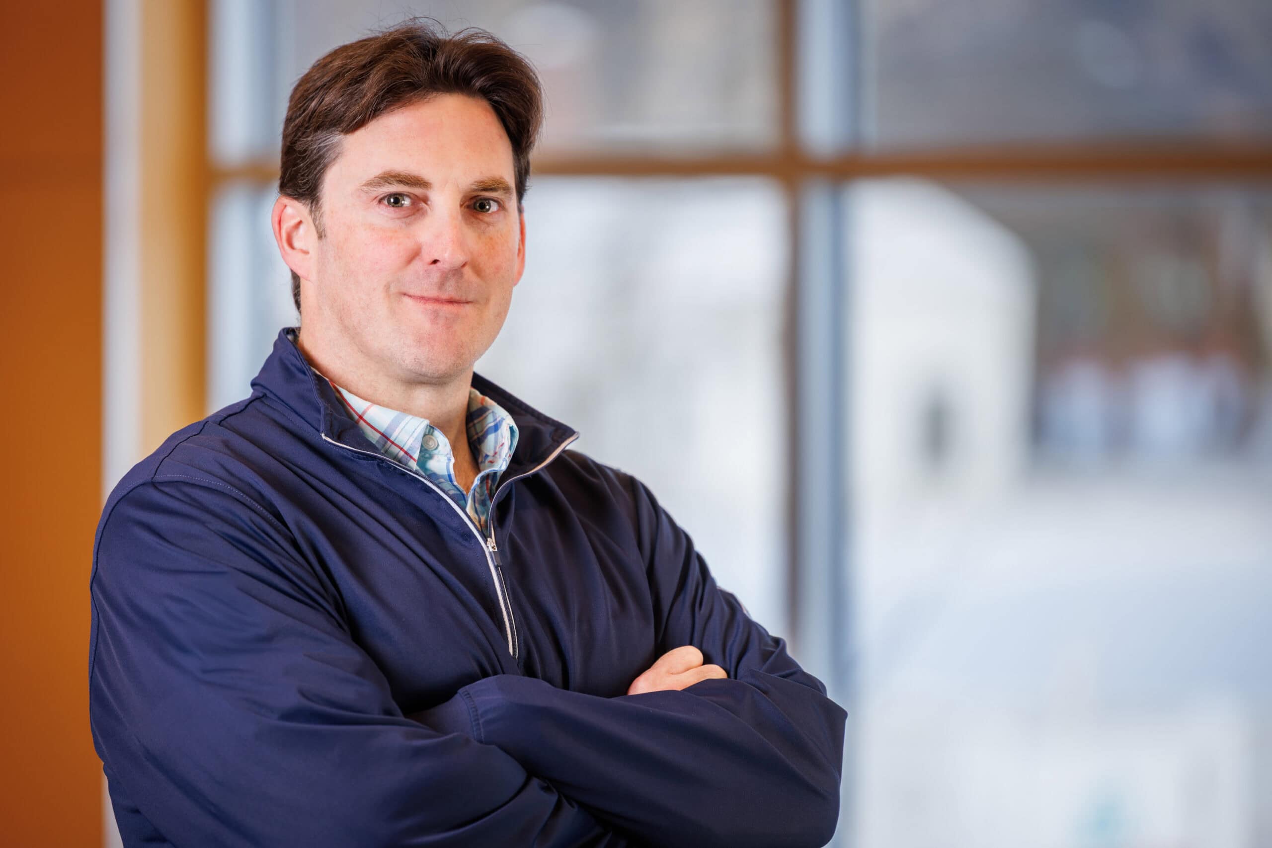 Bo McDougall, a man with short brown hair, is wearing a blue and pink checkered shirt with a navy blue jacket over top, smiling at the camera.