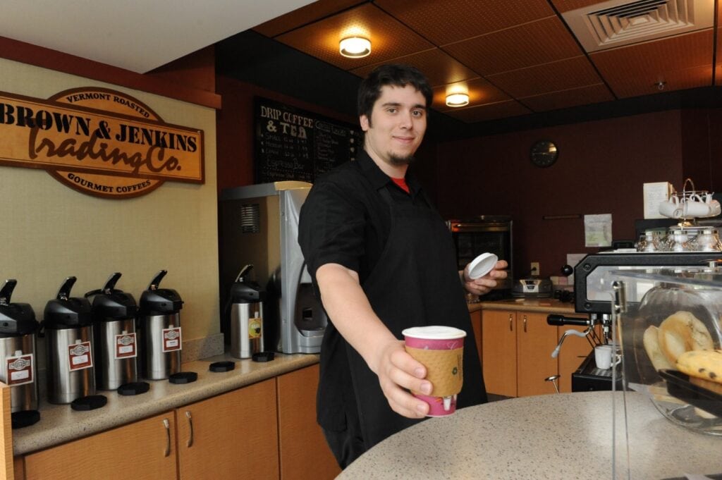 A young man with dark hair holds out a cup of coffee towards the camera on the Vermont State University Johnson campus.