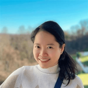 Woman with black hair and tan sweater smiles at the camera with a forest in the background.