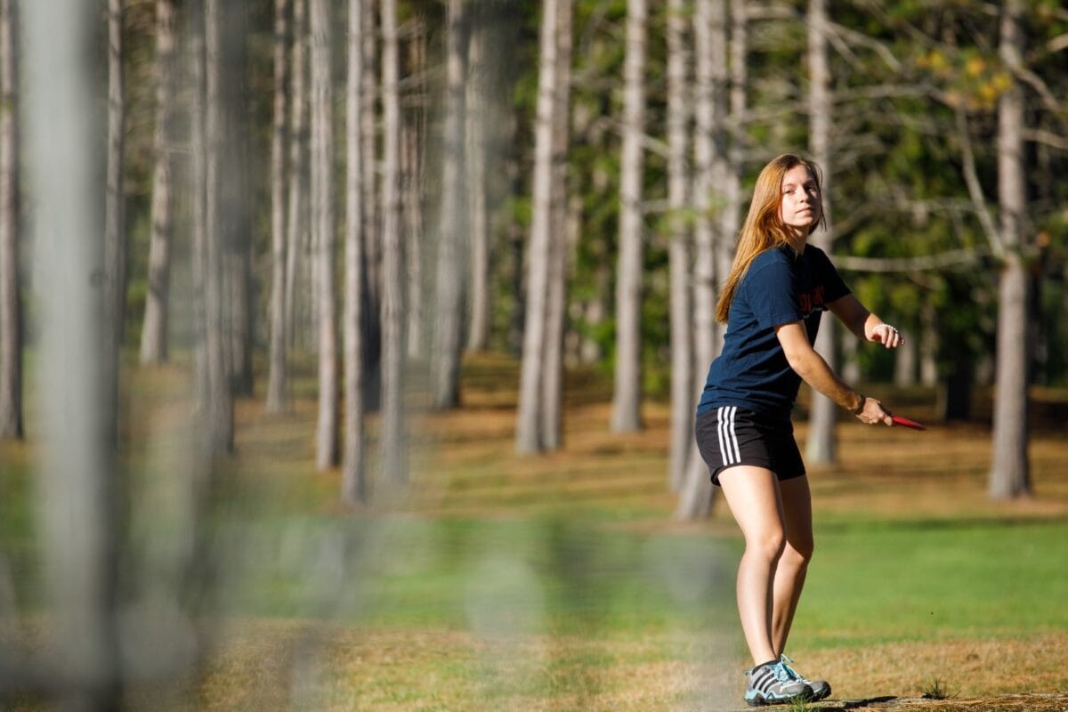 A young girl with long red hair throws a disc at a basket on the Vermont State University Johnson disc golf course.