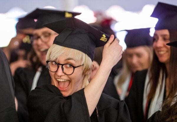 A student with short blond hair and glasses shouts with glee at the camera while shifting the tassel on their graduation cap at the Johnson campus commencement.