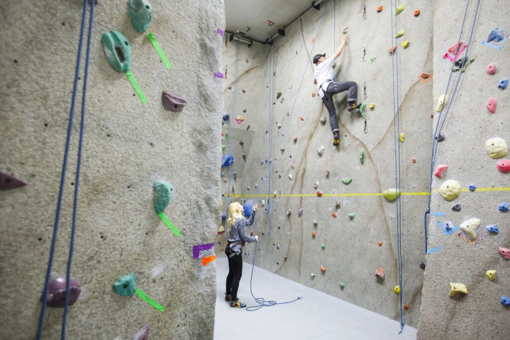 A young man climbs an indoor climbing wall while a woman belays him on the Vermont State University Lyndon campus.