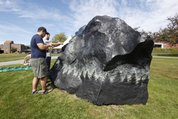 Two young men paining a giant black boulder on the Vermont State University Johnson campus.