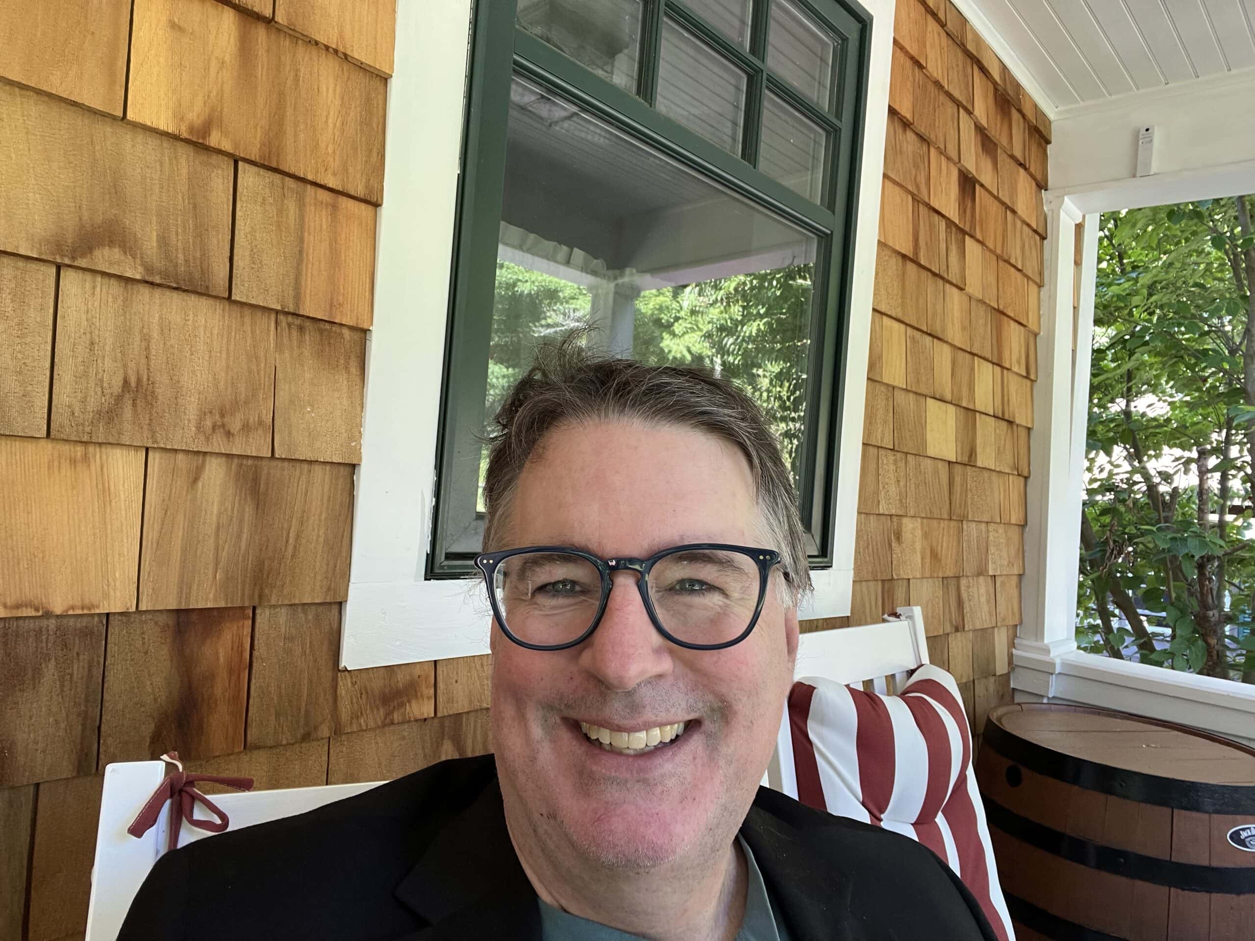 Scott Roper sitting in a white chair on a porch and smiling directly at the camera