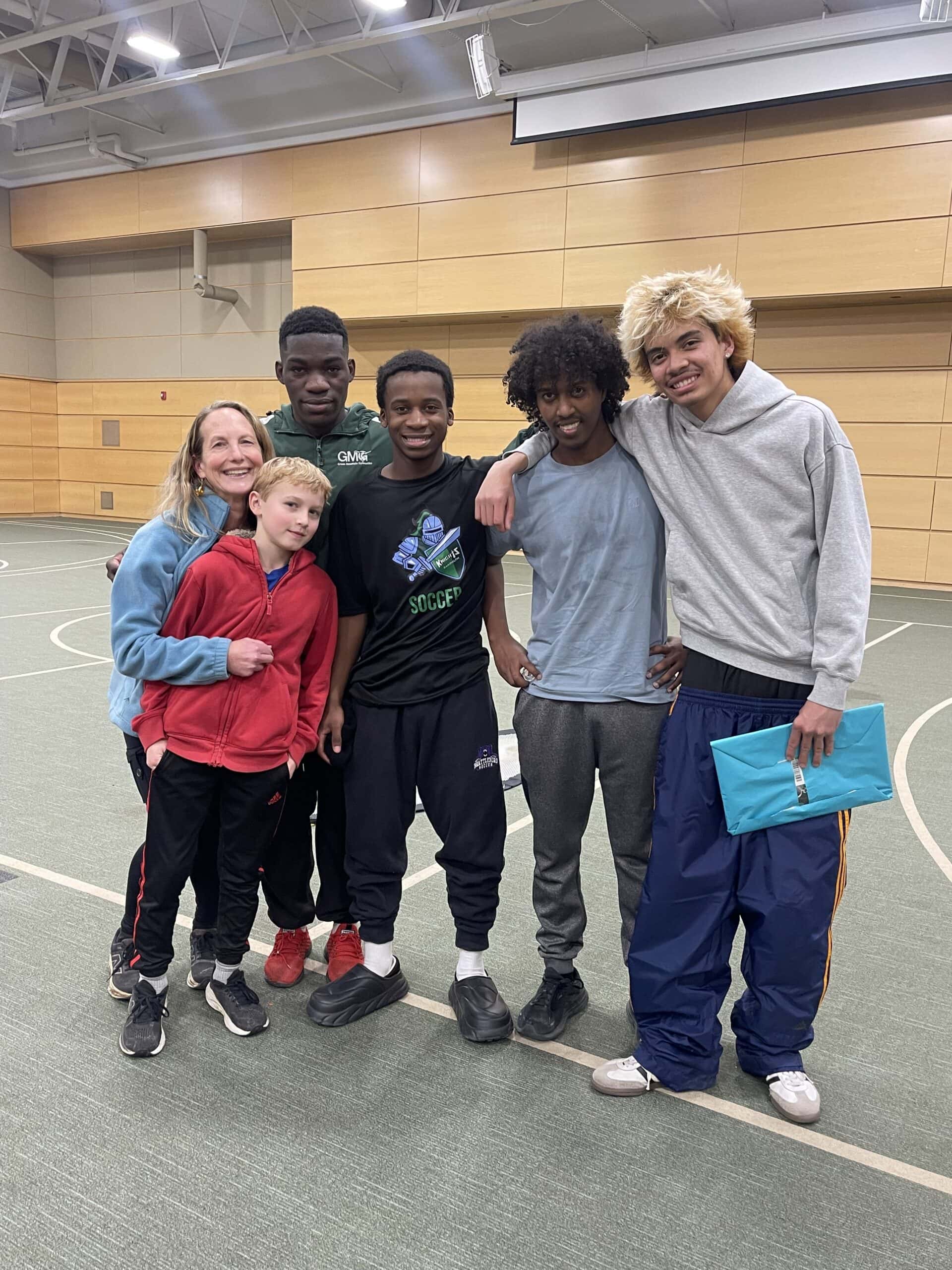 A group of six people stand together in an indoor gymnasium with a green sports floor and wooden-paneled walls. They are posed closely with arms around one another. The group includes a mix of children and teens dressed in casual athletic clothing—sweatshirts, track pants, sneakers. One person on the right holds a bright blue folder. The gym’s high ceiling, lights, and part of a basketball hoop are visible in the background.