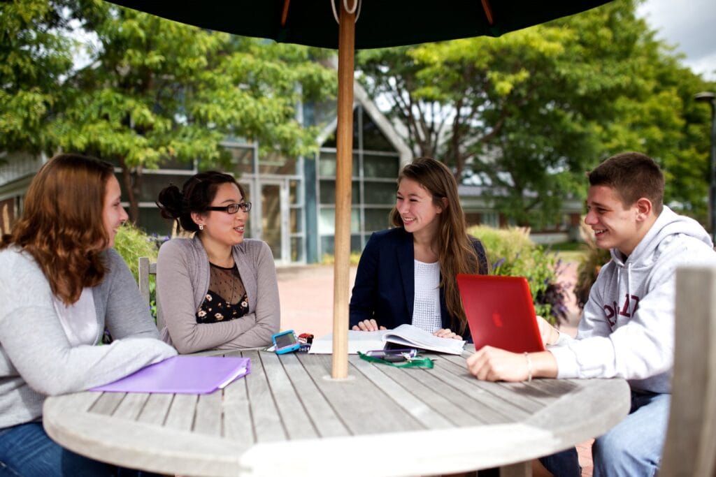 Friends gather outside at a picnic table under an umbrella on the Vermont State University Randolph campus.