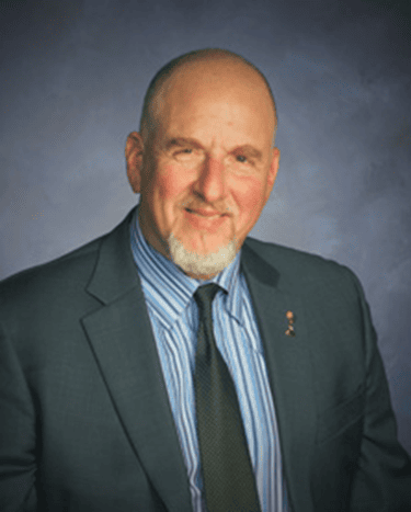 A man with a grey beard wearing a blue suit and striped shirt smiles at the camera in front of a grey background