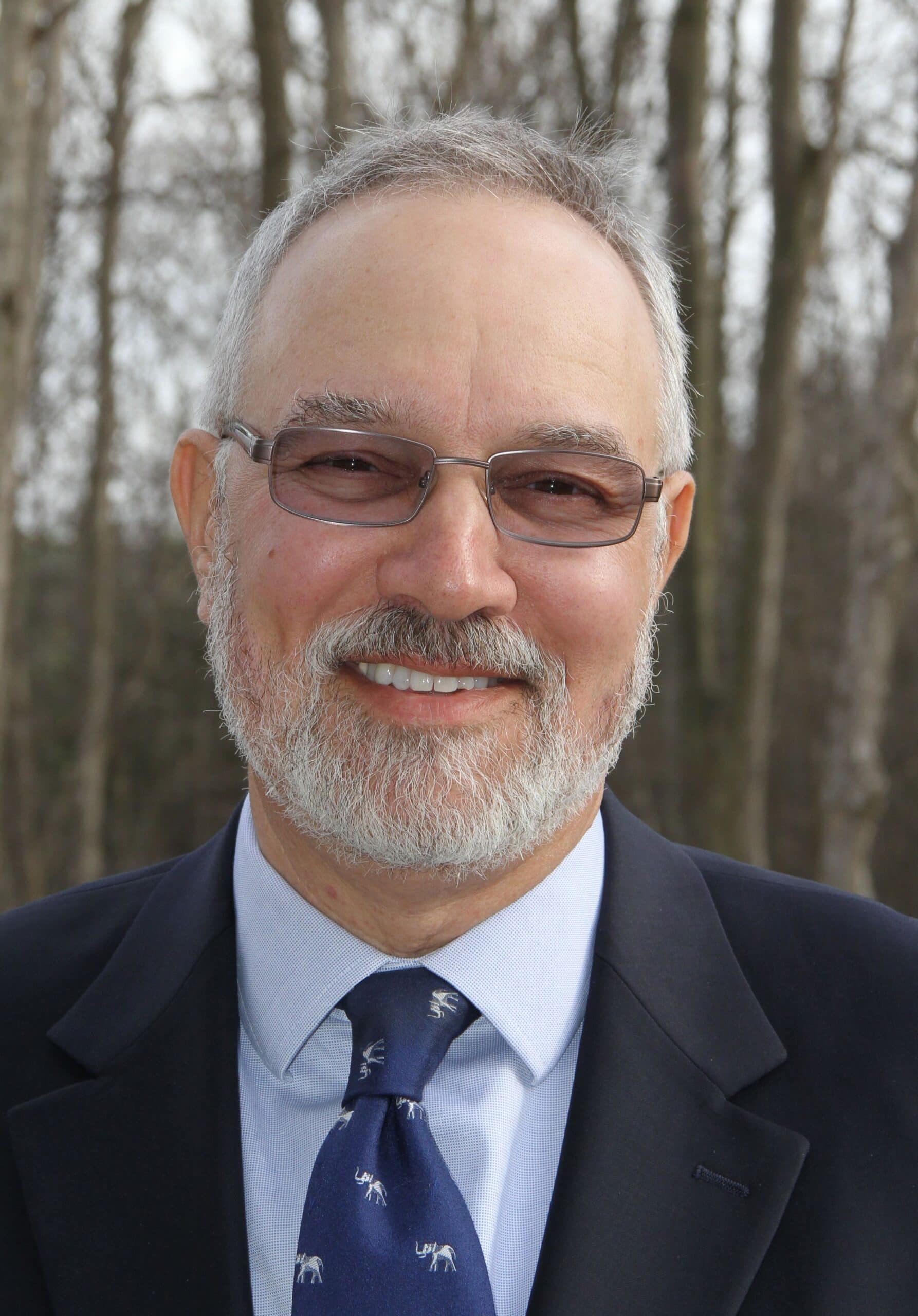 Man in a suite and tie, smiles at the camera with trees in the background.
