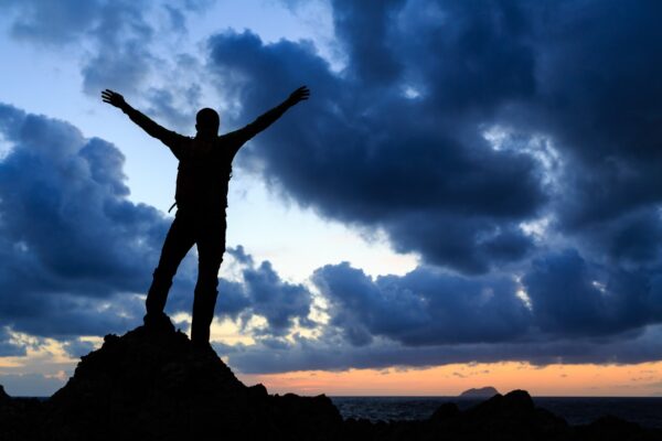 A person standing at the top of a mountain at dawn with his arms raised in triumph.