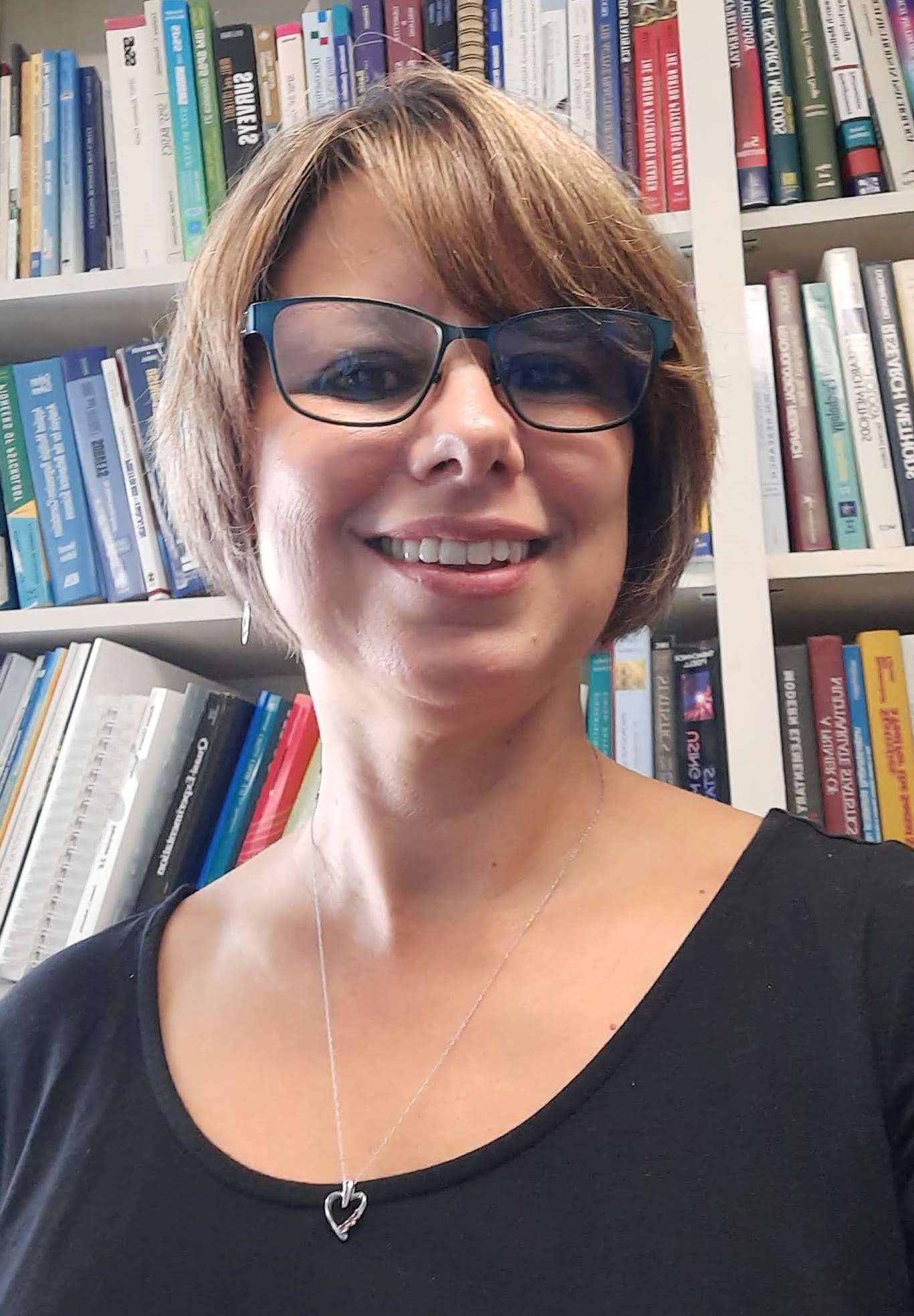 Woman in black shirt and short blonde hair with blue glasses, smiles at the camera with a bookcase in the background