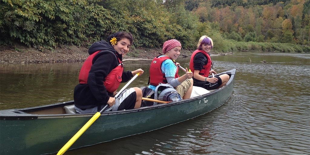 Three friends in a canoe turn and smile at the camera as they paddle down a river.