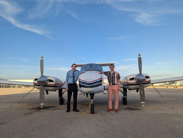 Two people stand in the front of an airplane on a runway.