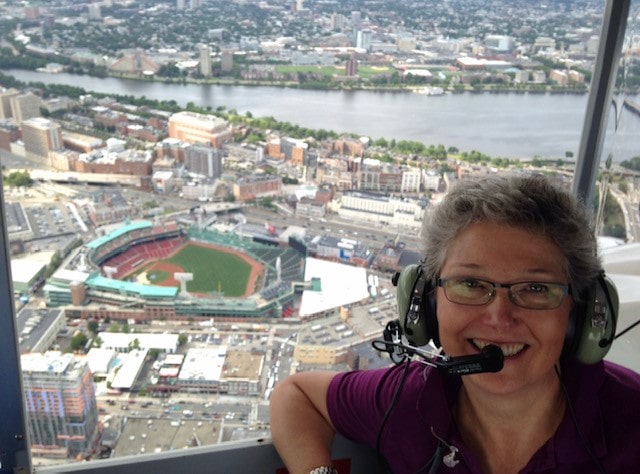 A woman wearing a headset smiles at the camera from inside a plane flying over a baseball stadium/