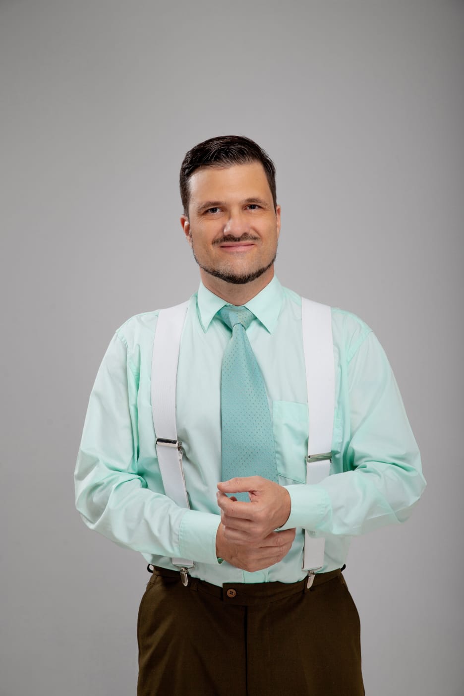 Man in a light blue shirt and tie smiles at the camera with a grey background.
