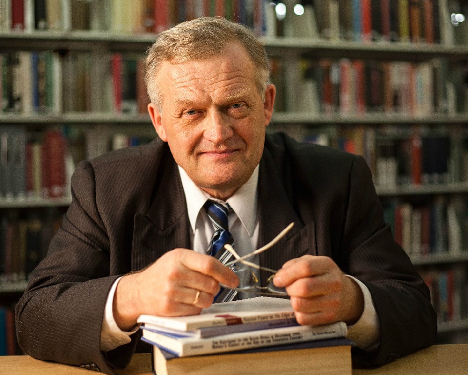 Man in a brown suit in a library smiles at the camera.