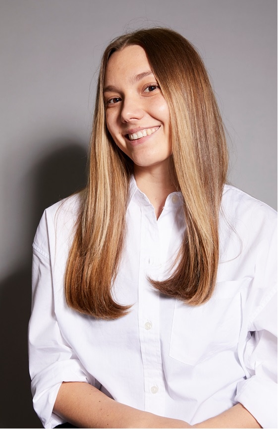 A young woman with long brown hair smiles sheepishly at the camera and wears a white button-up shirt.
