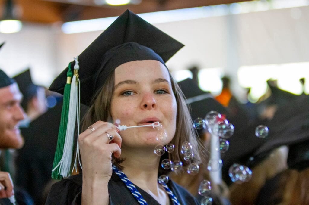 A Vermont State University graduate blowing bubbles at the camera in commencement regalia. 