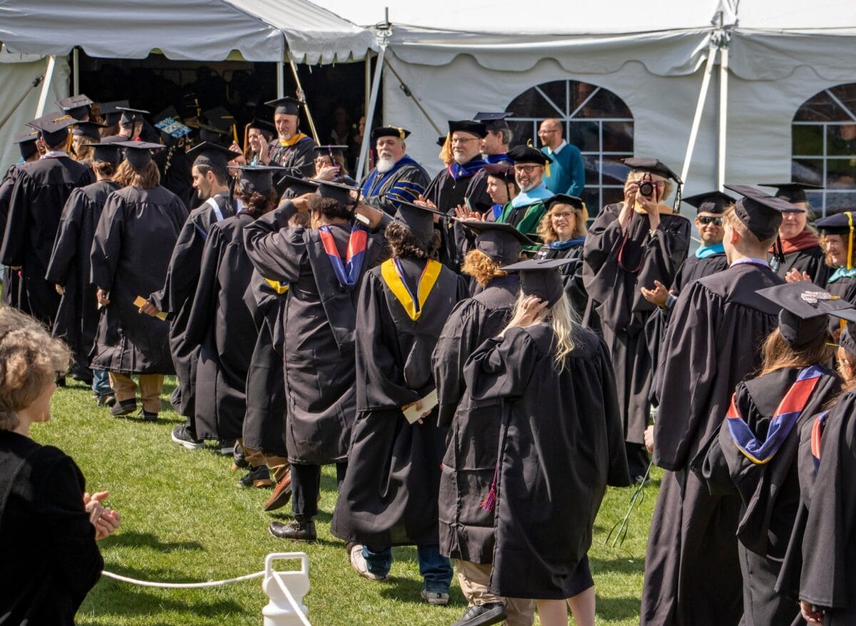 A group of graduating seniors walk into a tent on a sunny day and people cheer around them.
