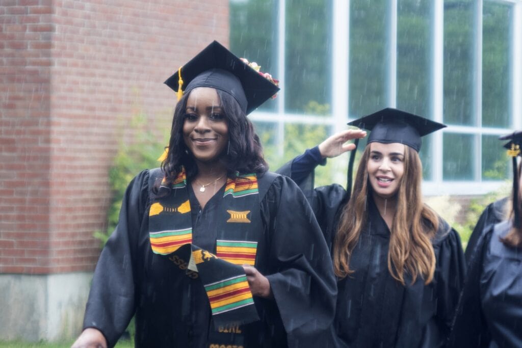 A Vermont State University graduating senior smiles at the camera as she walks through the rain in graduation regalia.