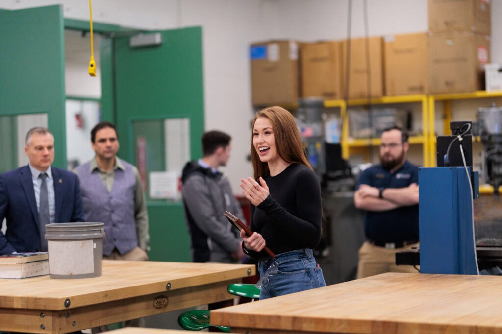 Sophia Rivera, a young woman with long brown hair wearing a black long sleeved shirt and blue jeans, smiling at a group of legislators in an engineering lab.