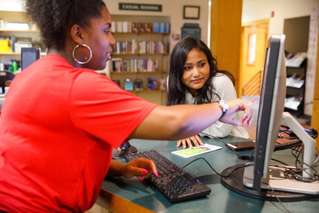 A young woman points at a computer monitor while another leans forward and looks.