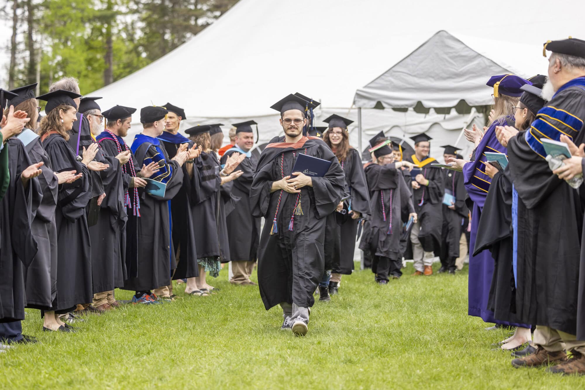 A line of graduates in caps and gowns walking down a grassy path with faculty and staff members on either side clapping.